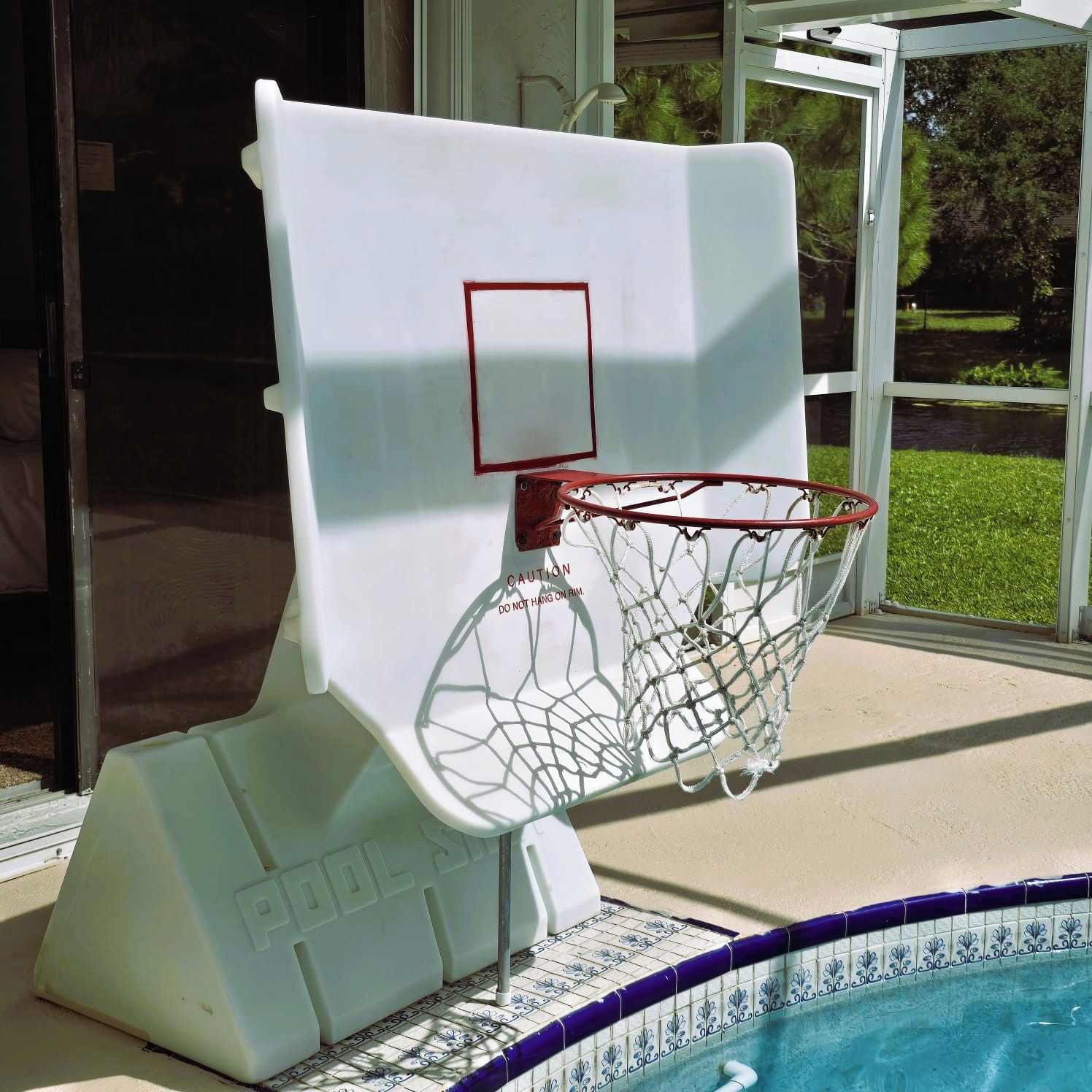 En basketkorg placerad vid poolkanten, tillsammans med en varningsskylt, en poolkant klädd med kakel och en utsikt över grönskan genom en glasdörr.