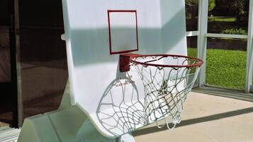 En basketkorg placerad vid poolkanten, tillsammans med en varningsskylt, en poolkant klädd med kakel och en utsikt över grönskan genom en glasdörr.