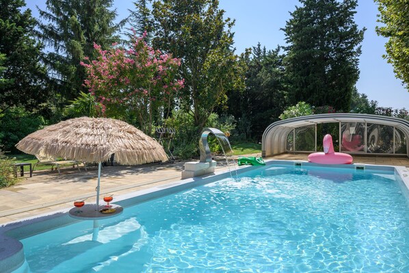 Une piscine agrémentée d’un parasol en chaume, d’un élément décoratif aquatique et d’une structure de serre.