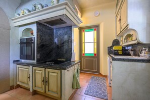 Eine Küche mit Backofen, Herd und Spüle. Es gibt ein Fenster mit Buntglas, und eine Tür mit dekorativer Klinke.