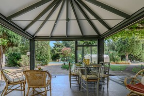 A covered patio with a table set for dining, surrounded by wicker chairs, and a view of a garden and pool area. - Reflet De Provence - Proche d'Avignon (Le Thor)