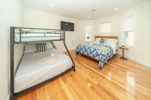 Quarto com beliche, cama individual, piso de madeira, televisão e janela com persianas.