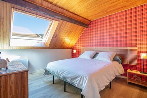 Ein Schlafzimmer mit schrägverdeckter Decke, einem großen Fenster, einem Bett mit Kissen, einem Nachttisch und einer Wand mit Karomuster.