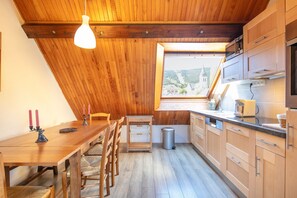 A kitchen with wooden cabinets, a dining table with chairs, a hanging light fixture, and a window with a view of a church and hills. - La Moucherolle - Station de ski accessible à pied (Méaudre)
