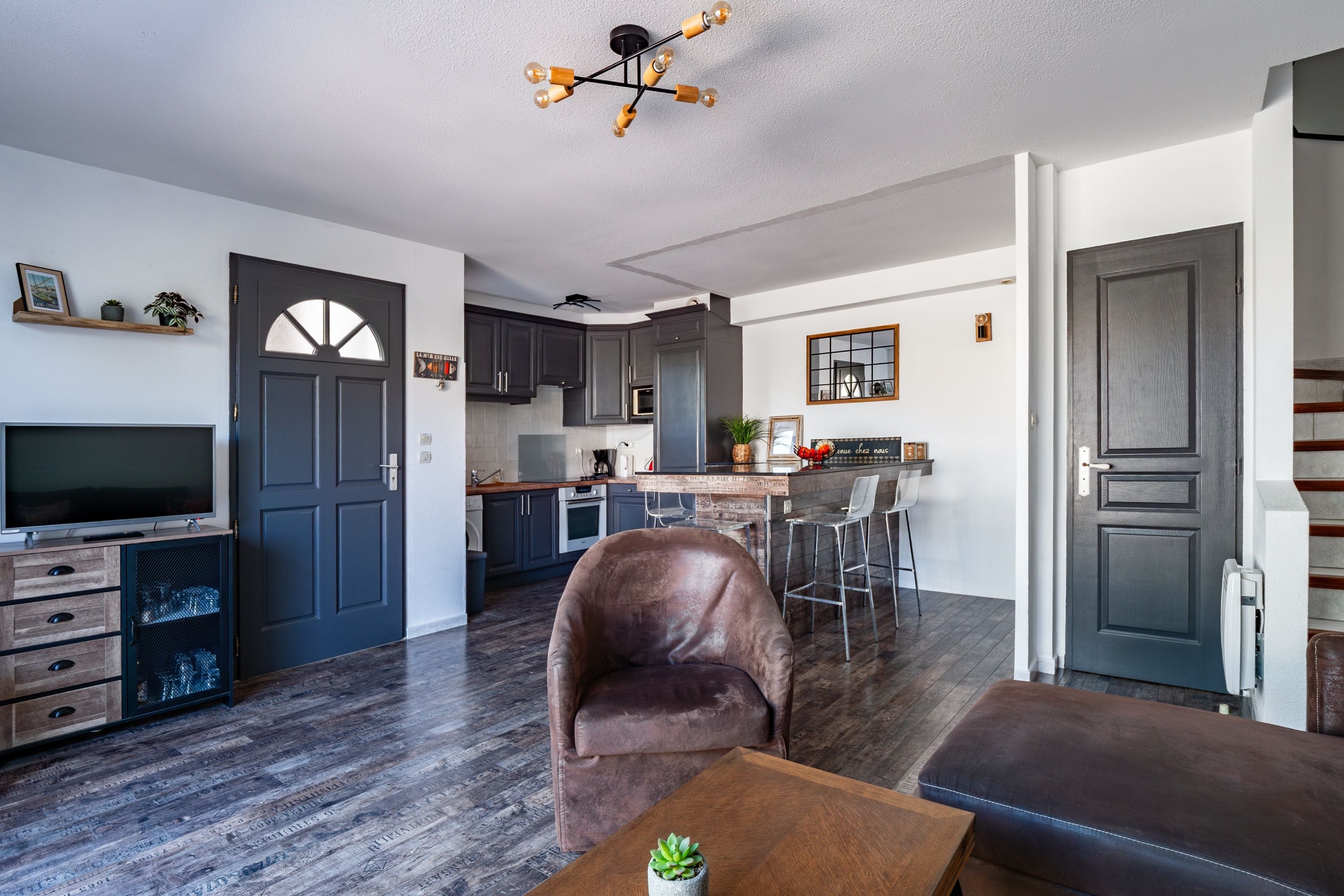 A modern kitchen with dark cabinets, a dining area with a leather chair, and a TV in the living room.