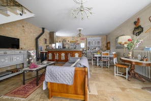 A spacious living room with a stone wall, a wooden sofa, a television, and a kitchen area in the background. - Le Coin Rétro - Proche d'Avignon (Le Thor)