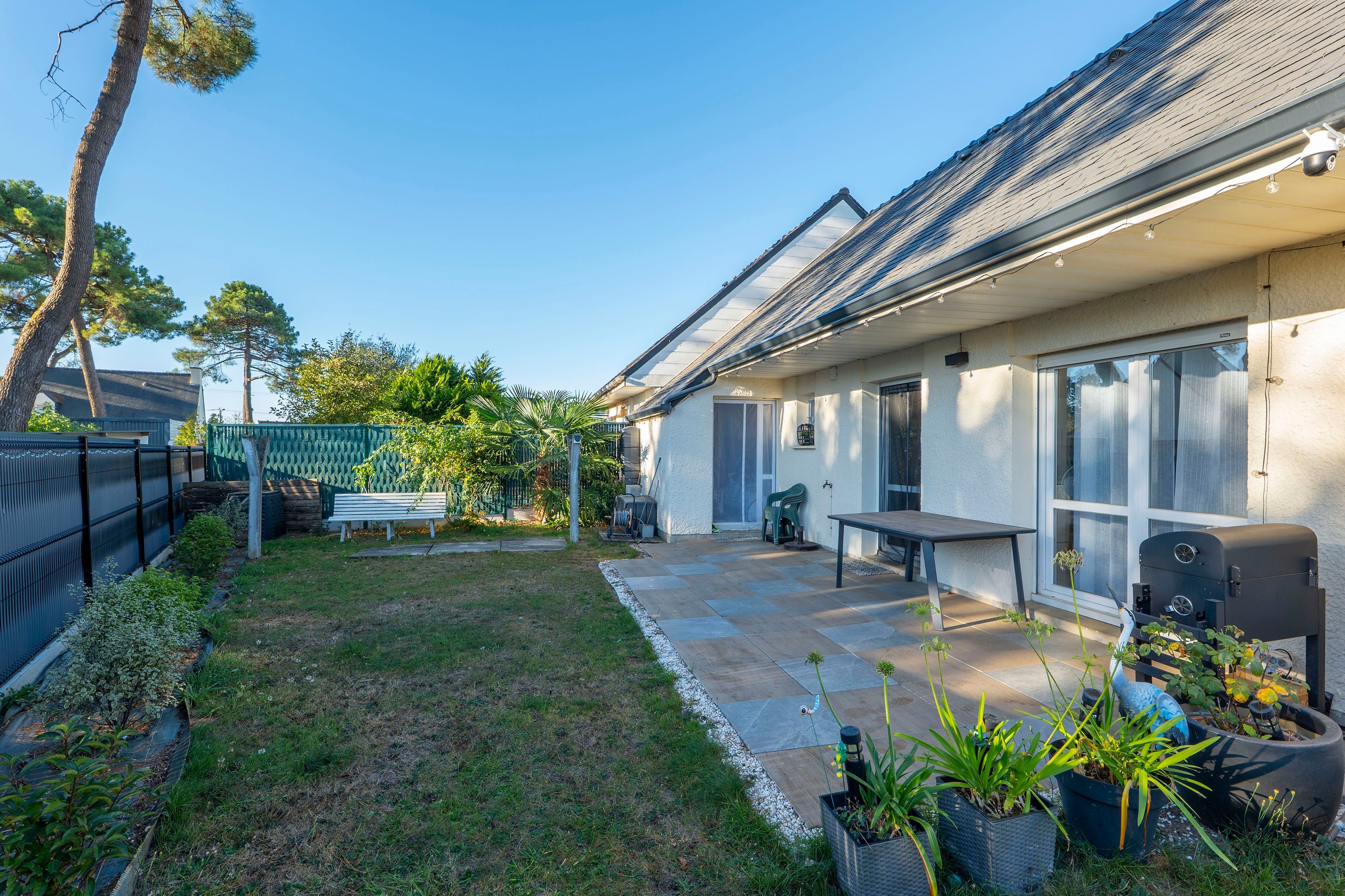 Un jardin à l’arrière d’une maison, avec une terrasse, un barbecue et des plantes en pot.