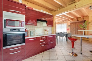 A modern kitchen with red cabinetry, a microwave, an oven, and a dining area with a table and chairs. - Chalet Caribou - Pour 6 avec terrasse (Ventron)