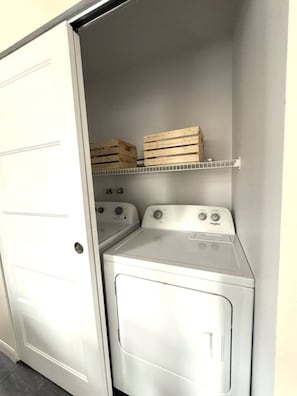 A laundry room with two stacked washers and dryers, wooden crates on a shelf, and a white door. - The hideaway of Limoilou, a cozy little urban nest (Québec)