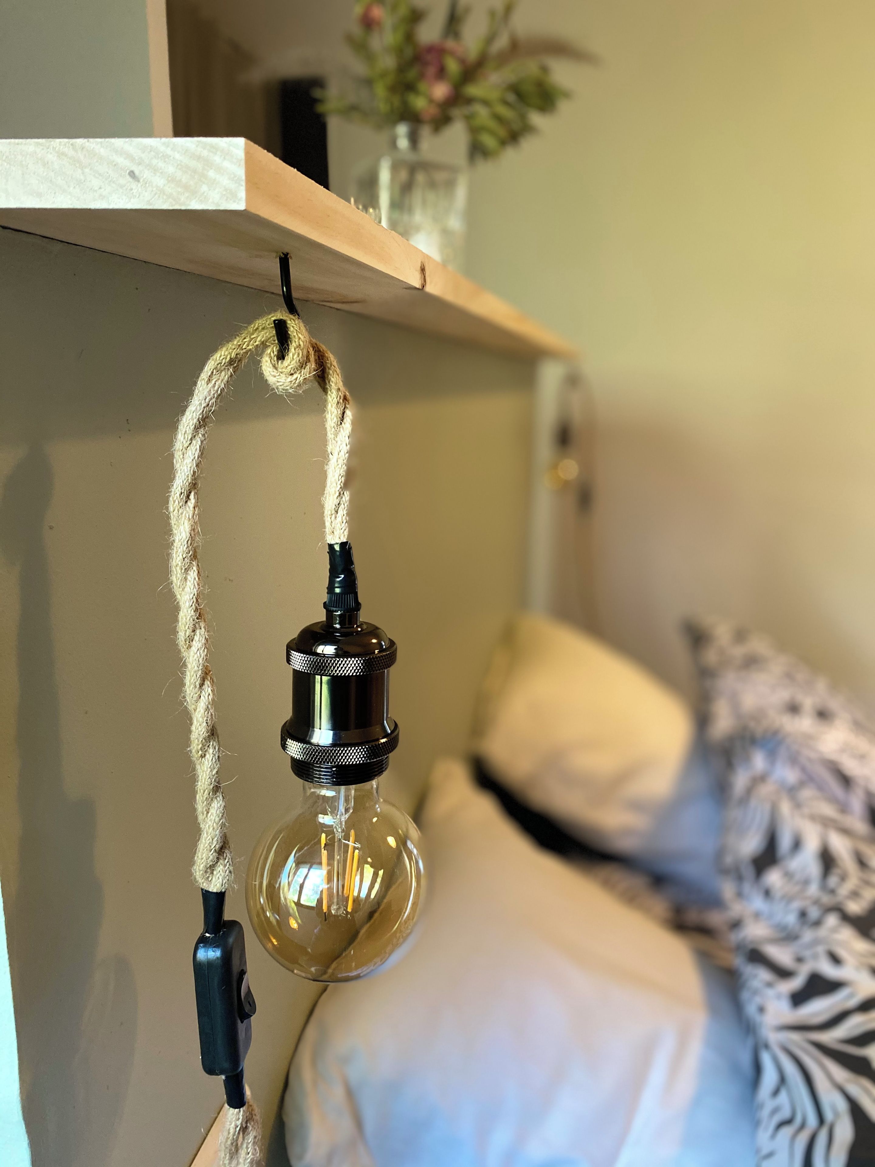 A hanging light fixture with a rope, a bed with white bedding, and a shelf with a vase of flowers.