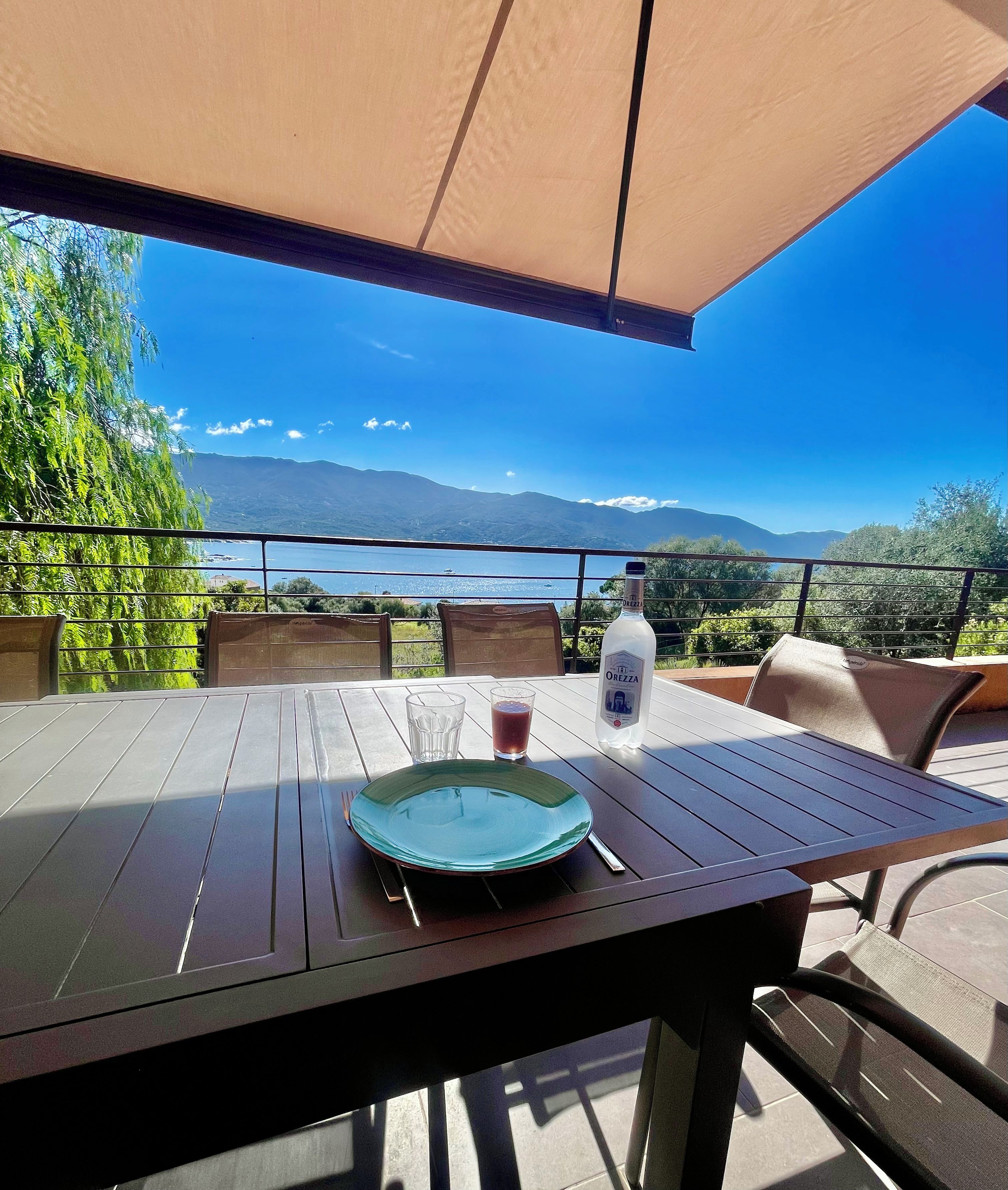 Une table avec une bouteille de liqueur OREZZA, un verre et une assiette, placée sur une terrasse extérieure d’où l’on peut admirer un lac et des montagnes.