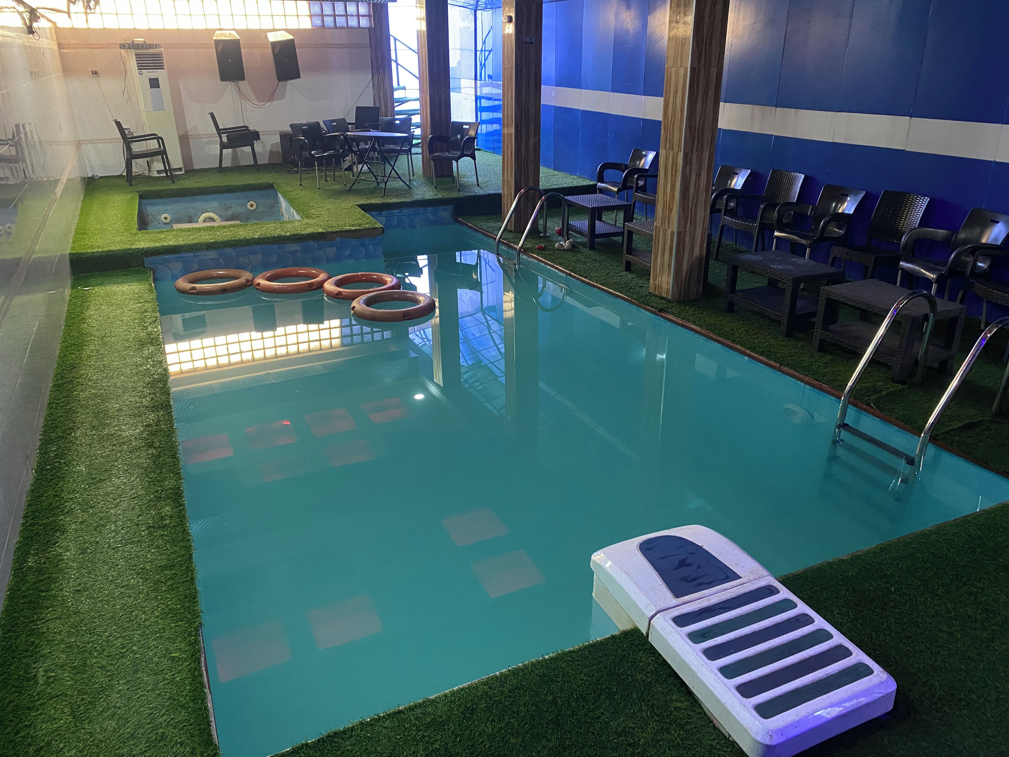 An indoor swimming pool with artificial grass borders, a lounge area with chairs, and a poolside table.