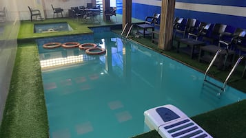 An indoor swimming pool with artificial grass borders, a lounge area with chairs, and a poolside table.