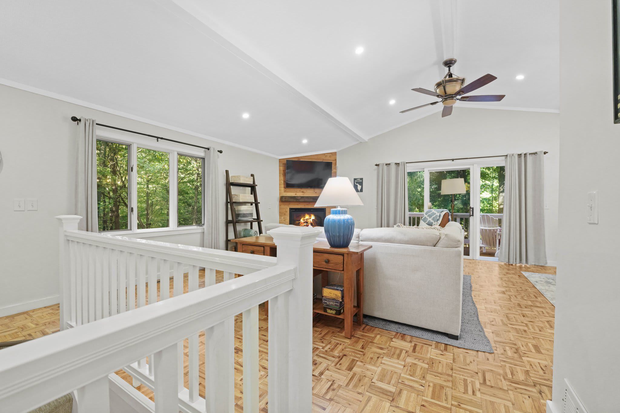 A spacious living room with a fireplace, a ceiling fan, and large windows.
