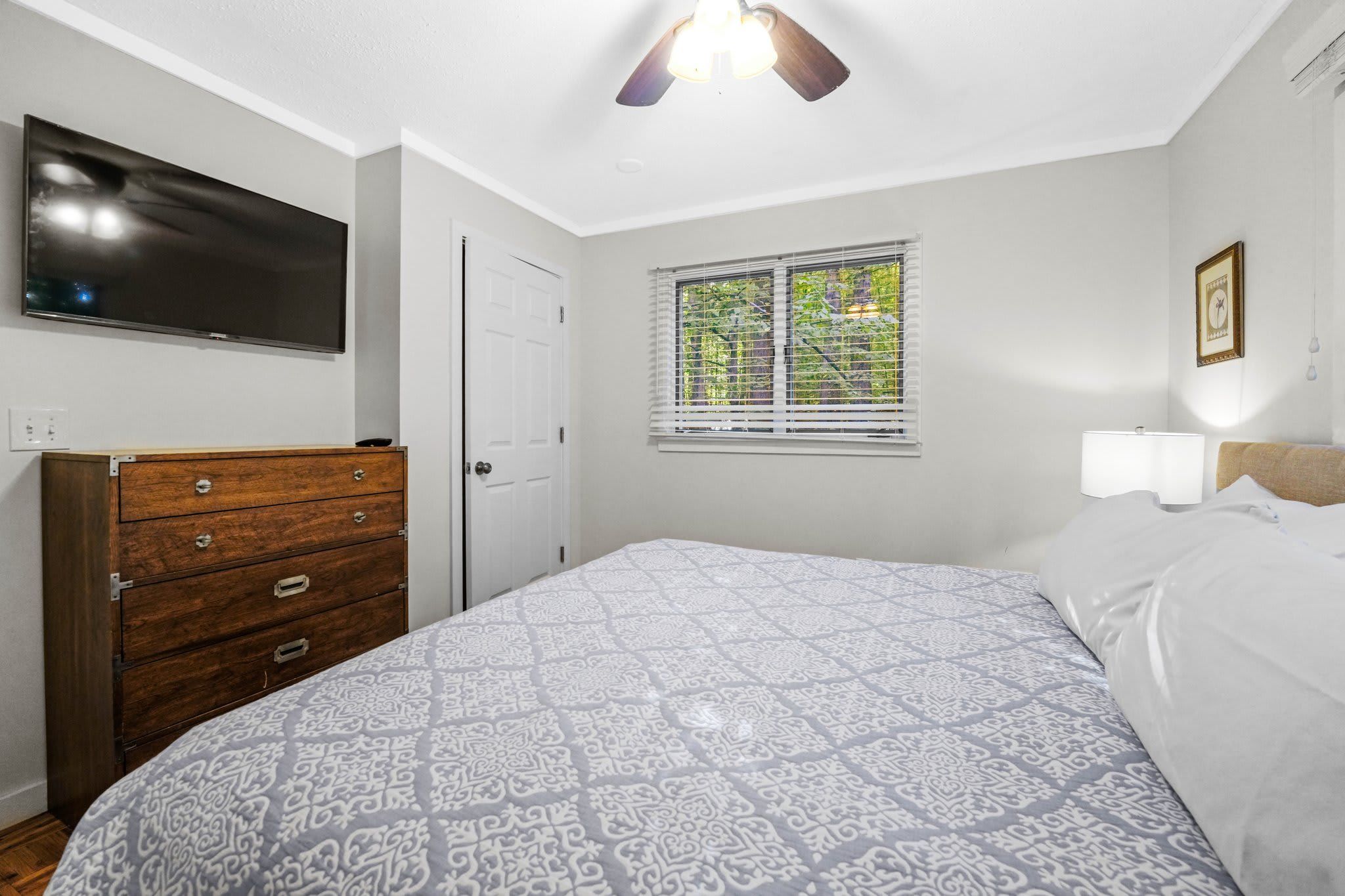 A bedroom with a bed, a dresser, a television, a window with blinds, and a ceiling fan.