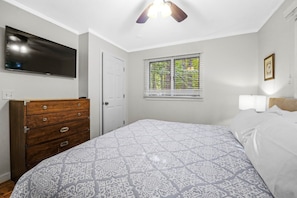 A bedroom with a bed, a dresser, a television, a window with blinds, and a ceiling fan.