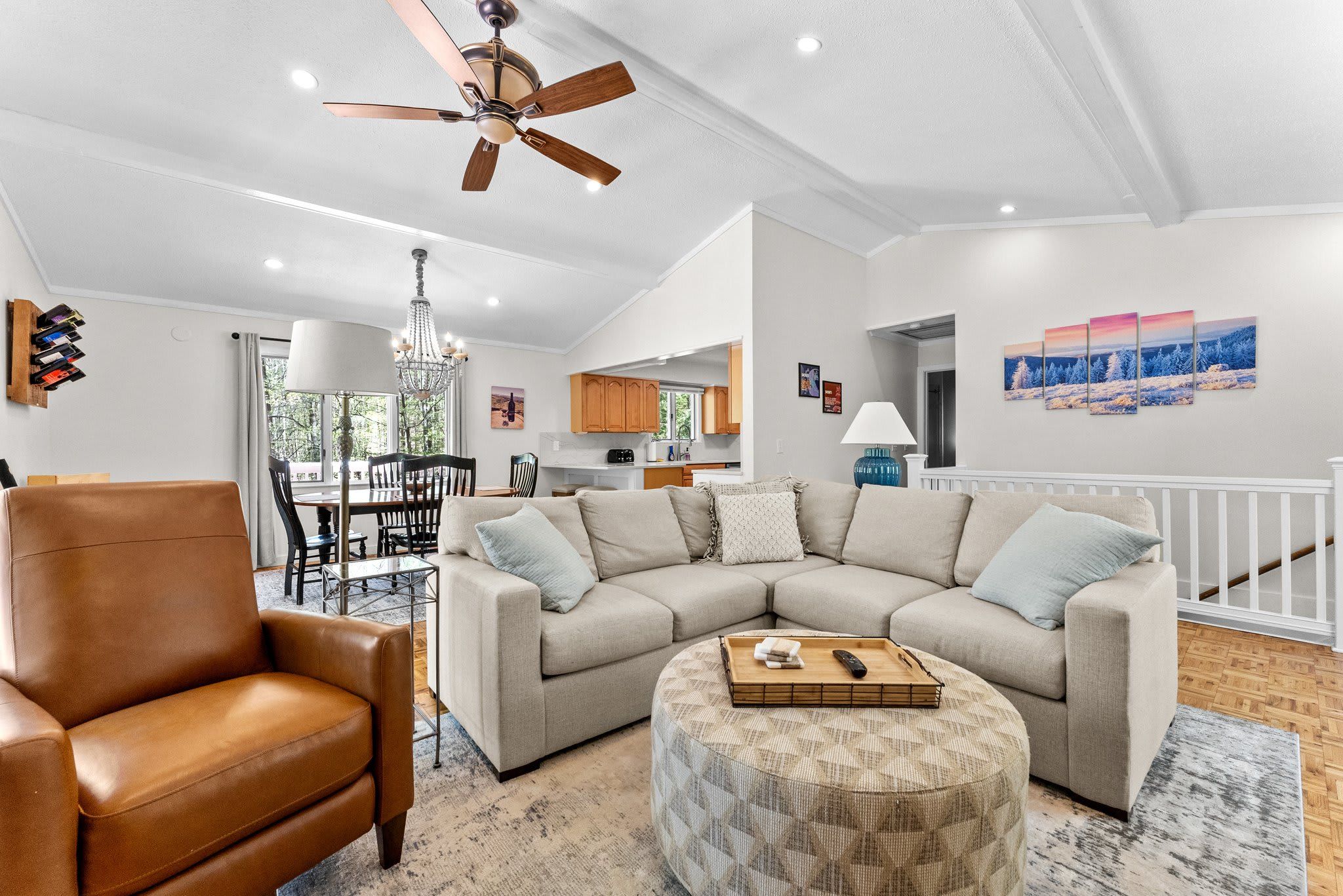 A modern living room with a leather armchair, a sectional sofa, and a round ottoman. There is a ceiling fan, a chandelier, and a dining area visible in the background.