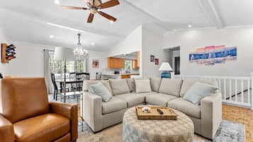 A modern living room with a leather armchair, a sectional sofa, and a round ottoman. There is a ceiling fan, a chandelier, and a dining area visible in the background.
