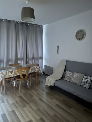 Un saló modern amb zona de menjador, un sofà gris amb coixins estampats, un rellotge de paret i una làmpada penjant.