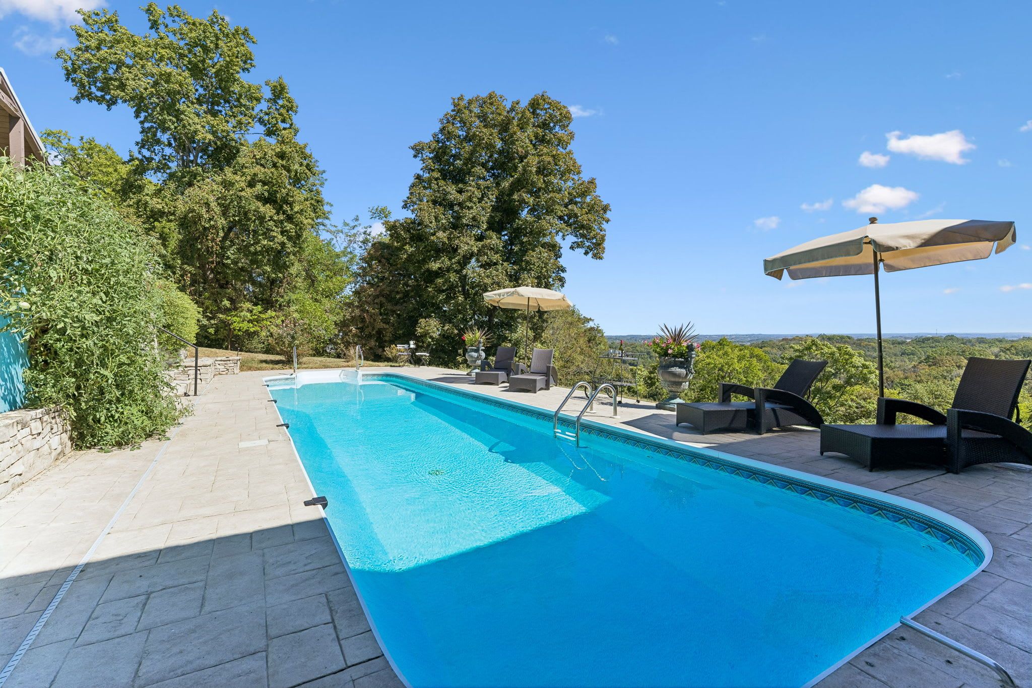 A rectangular swimming pool with a tiled deck, surrounded by lounge chairs and umbrellas, set against a backdrop of trees and a clear sky.