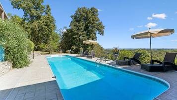 A rectangular swimming pool with a tiled deck, surrounded by lounge chairs and umbrellas, set against a backdrop of trees and a clear sky.