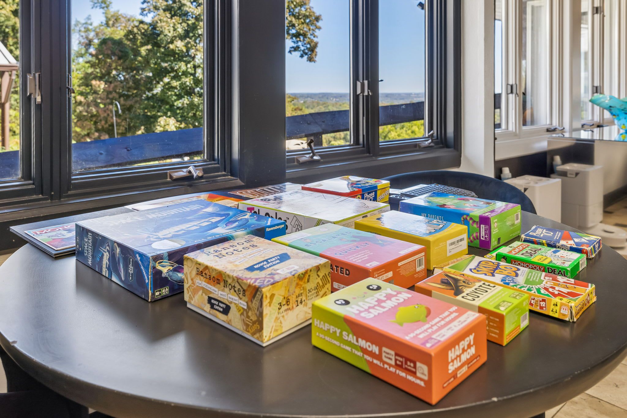 A table with various board games and game boxes, including Monopoly and Scrabble, in a room with large windows and a view of the outdoors.
