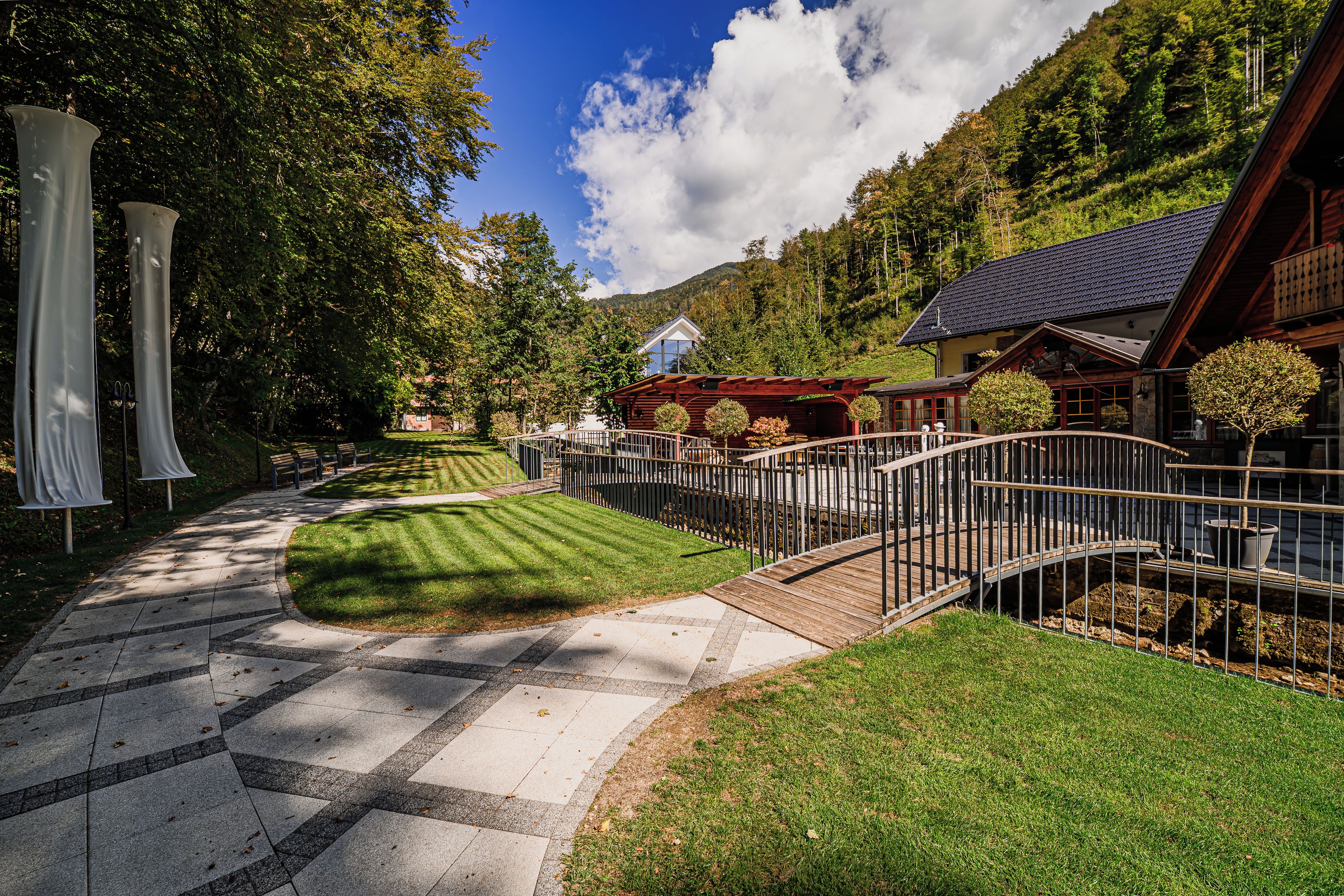 A well-maintained garden pathway leading to a wooden building with a sloped roof, surrounded by lush greenery and trees.