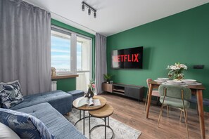 Un salon moderne avec un mur d’accent vert, un grand téléviseur à écran plat affichant le logo « NETFLIX », un canapé, une table basse et un coin repas comprenant une chaise et un vase de fleurs.
