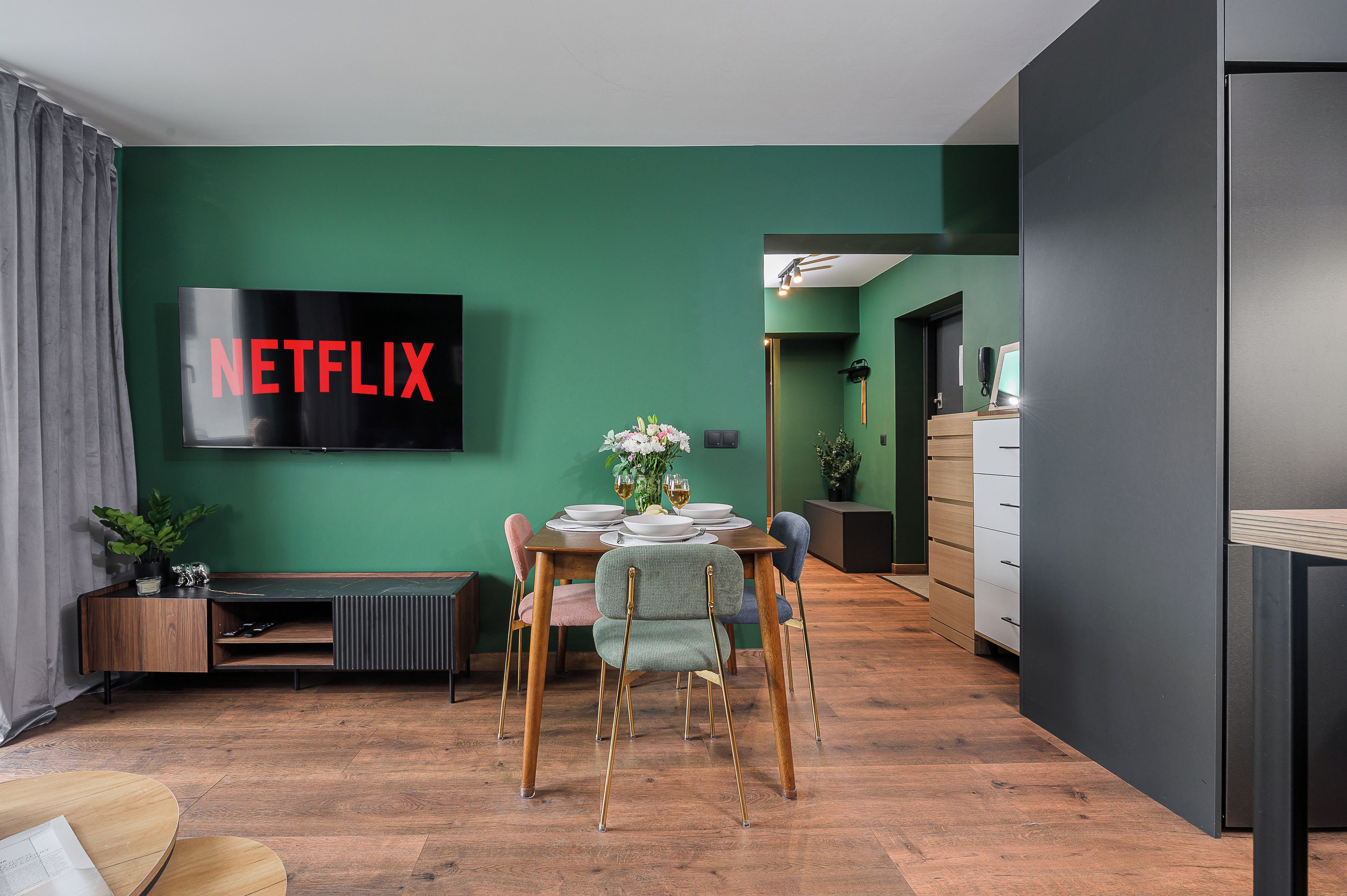 A modern living room with a dining area, a flat-screen TV mounted on a green wall, and a wooden floor.