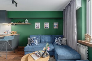 Ein modernes Wohnzimmer mit einer grünen Akzentwand, einem blauen Sofa mit gemusterten Kissen, einem kleinen Esstisch und einem Fenster mit Vorhängen.