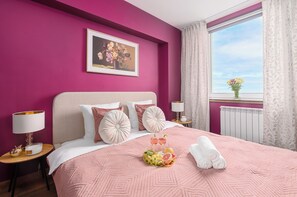 Ein Hotelzimmer mit einem großen Bett, einem Gemälde an der Wand, einem Fenster mit Vorhängen und einer Obstschale auf dem Bett.