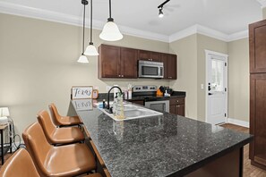 A kitchen with a granite countertop, brown leather bar stools, a stainless steel microwave, and pendant lighting. - Bring the group! Comfort-First Cottage Living (College Station)