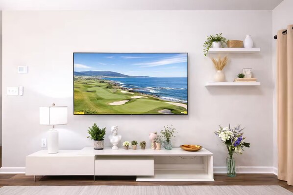 A modern living room with a flat-screen TV mounted on the wall, a white entertainment unit, and decorative items including plants and a vase. - Brand New 3BR Home - Sleeps 10 - Pool Access (Myrtle Beach)
