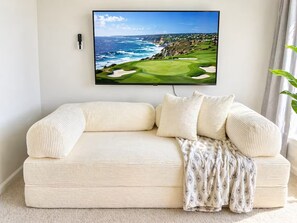 A living room with a large flat-screen TV mounted on the wall, a cream-colored sofa with multiple cushions, and a patterned throw blanket. - Brand New 3BR Home - Sleeps 10 - Pool Access (Myrtle Beach)