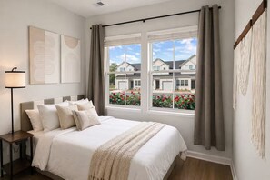 A bedroom with a bed, a nightstand, a lamp, and a view of houses through a window. - Brand New 3BR Home - Sleeps 10 - Pool Access (Myrtle Beach)