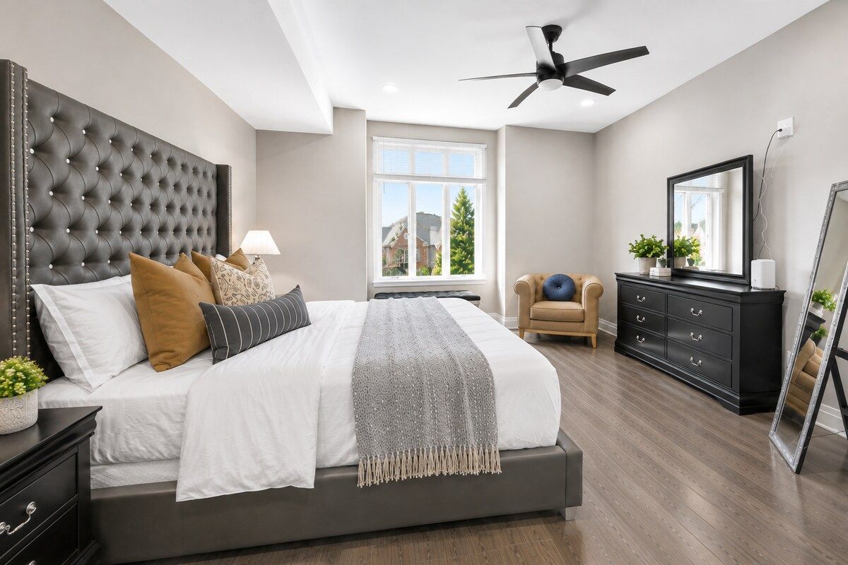 A bedroom with a tufted headboard, a bed with multiple pillows, a dresser, a chair, a mirror, and a ceiling fan.