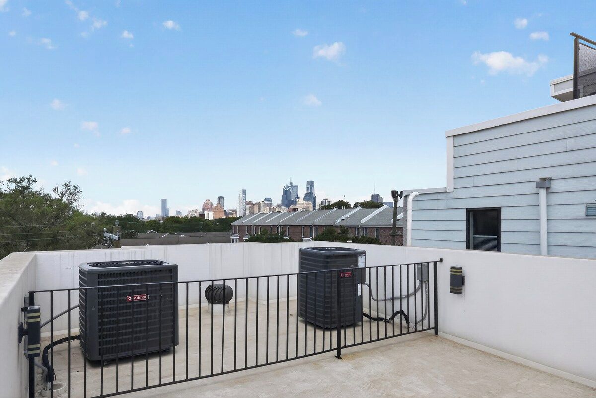 A rooftop terrace with two air conditioning units, a metal fence, and a city skyline in the background.