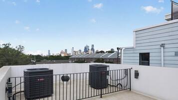 A rooftop terrace with two air conditioning units, a metal fence, and a city skyline in the background.