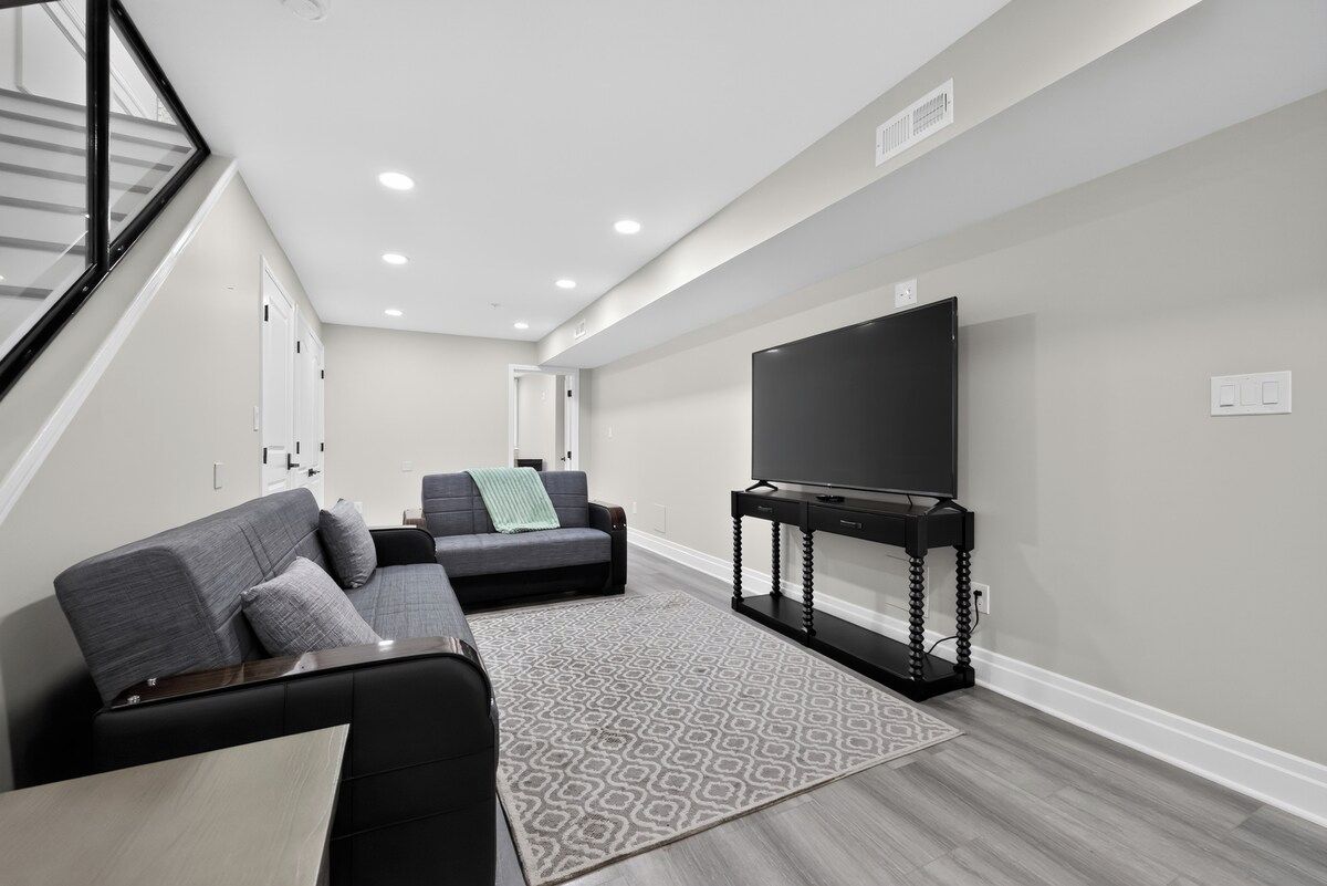 A modern living room with a sectional sofa, a flat-screen TV on a stand, and a staircase with a glass railing.