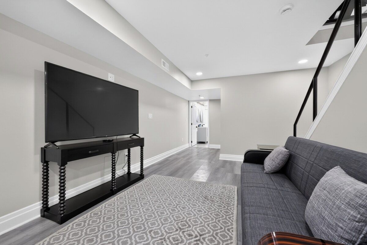 A modern living room with a flat-screen TV on a black stand, a grey sofa, and a patterned rug.