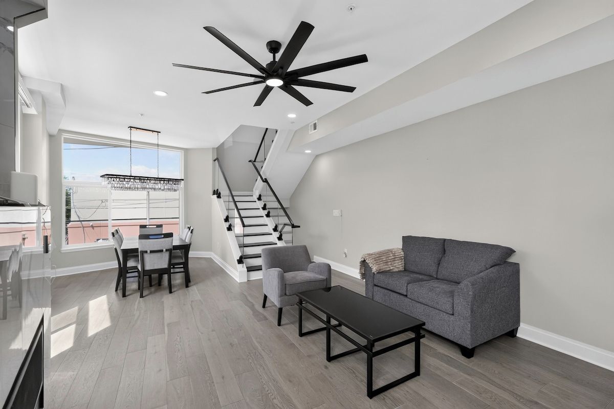 A modern living room with a staircase, a dining area, and a ceiling fan.