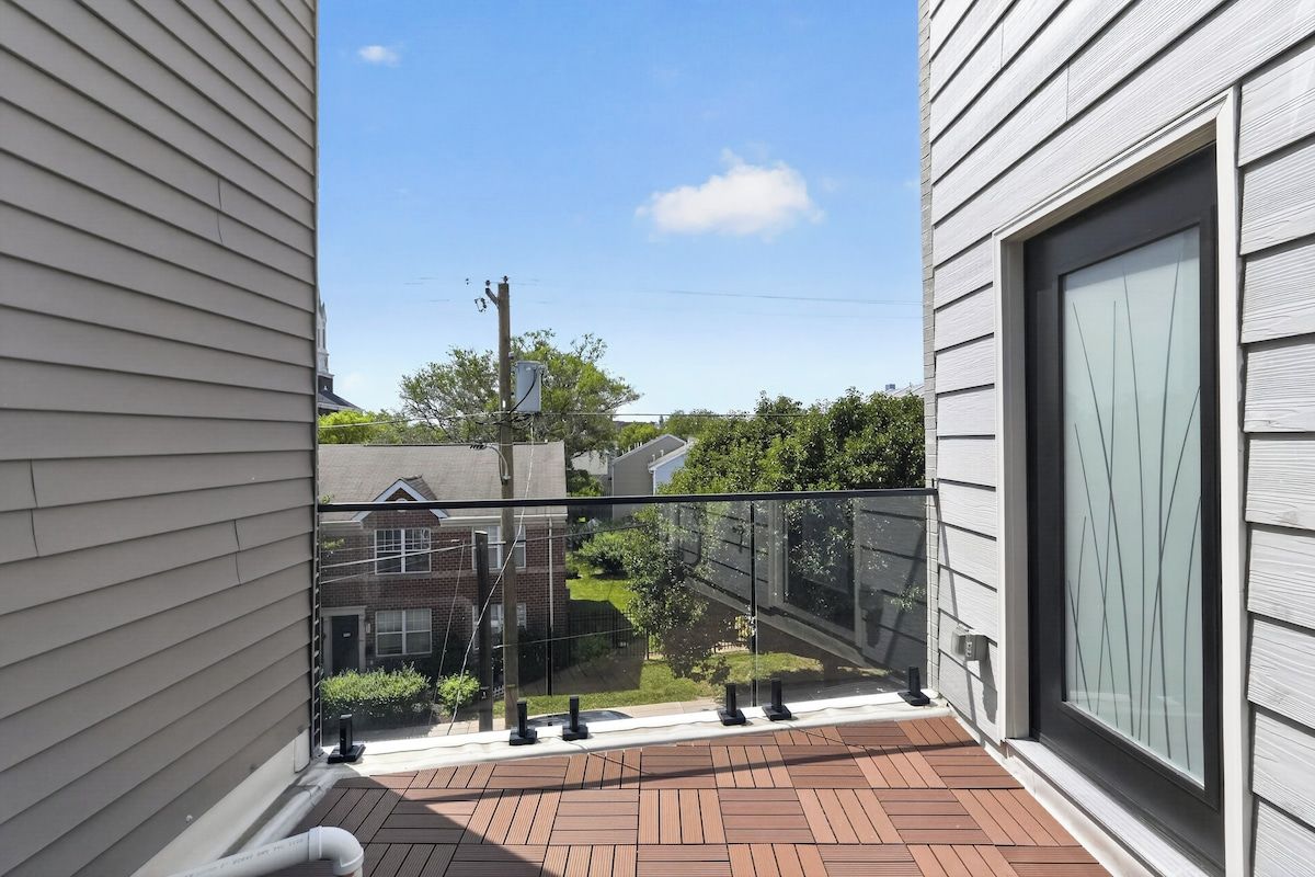 A balcony with a glass railing, a brick floor, and a view of a residential area with houses and trees.