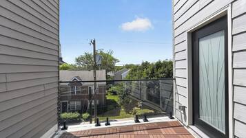 A balcony with a glass railing, a brick floor, and a view of a residential area with houses and trees.