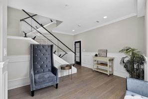 A spacious living area with a grand staircase, a tufted armchair, a small round table, and a potted plant. - Queen Village Grand | 3BR, Rooftop & Garage (Philadelphia)