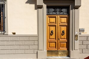 Une porte en bois à deux battants, surmontée d’un élément décoratif en métal, un mur en briques et une petite fenêtre avec une grille en métal.