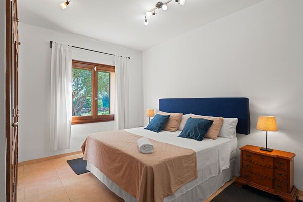 Ein Schlafzimmer mit einem Bett, einem Nachttisch, einer Lampe, einem Fenster mit Vorhängen und Blick auf Grünfläche.