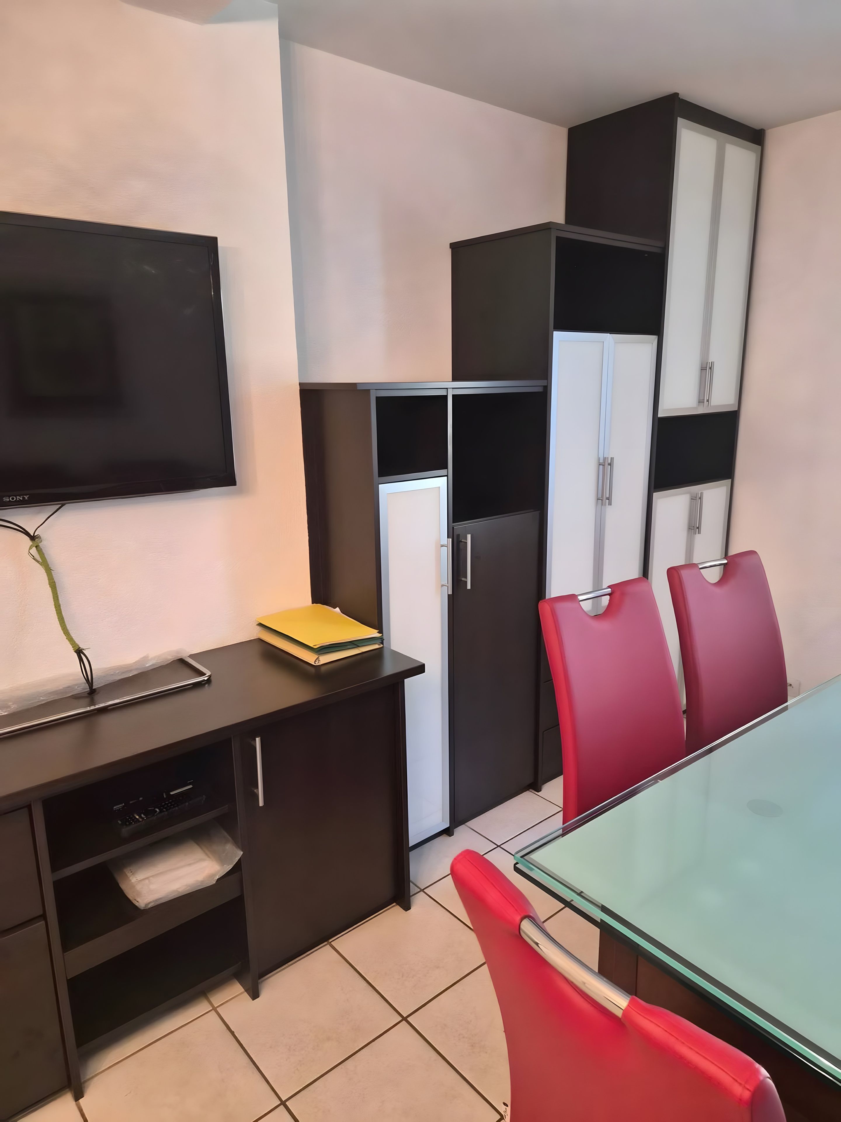 A modern kitchen with a flat-screen TV, a refrigerator, and a dining table with red chairs.
