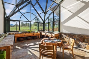 Un invernadero con una mesa y sillas de madera, ventanas amplias y vistas a un paisaje verde.