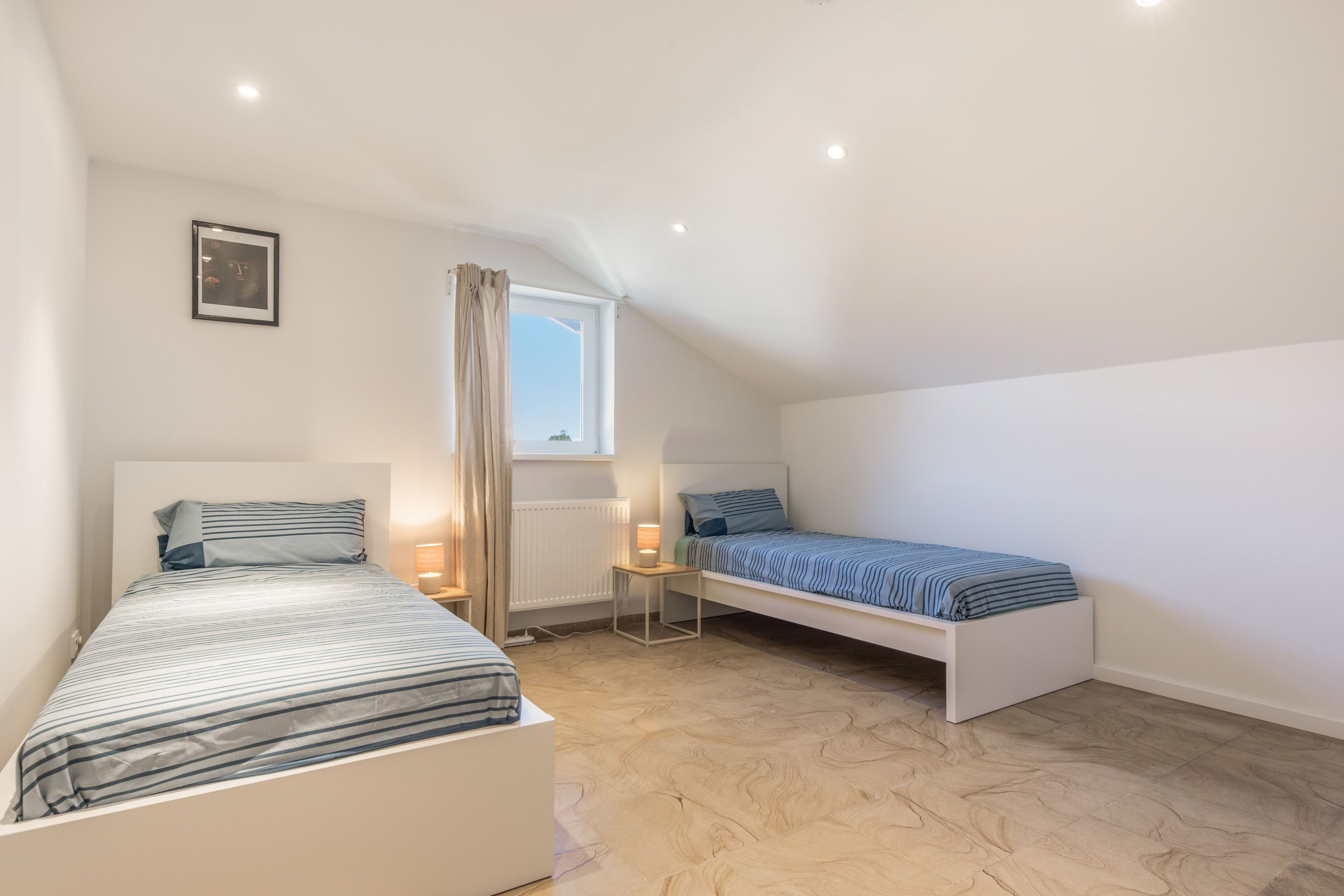 A bedroom with two beds, a window, a radiator, and a picture on the wall.