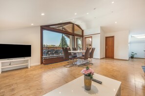 A spacious living room with a flat-screen TV, a dining area with chairs, and a large window offering a view of the outdoors.